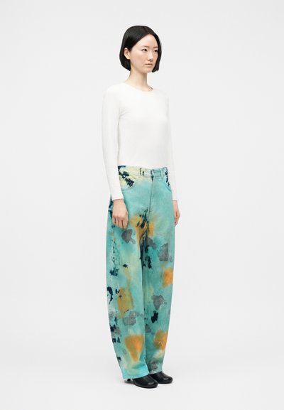 Γυναίκα με κοντά μαύρα μαλλιά φοράει λευκή μπλούζα με μακριά μανίκια και φαρδιά τιρκουάζ παντελόνια με πορτοκαλί και μαύρα σχέδια που μοιάζουν με πιτσιλιές μπογιάς.