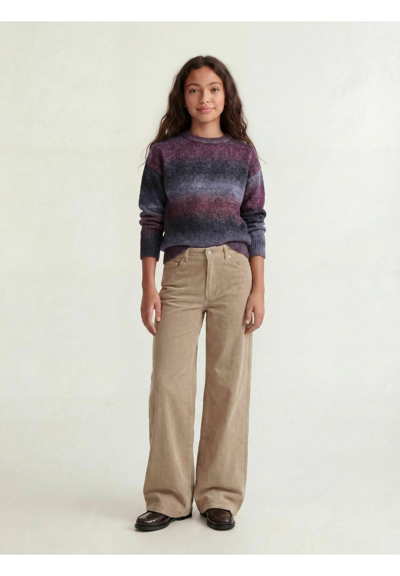 Jeune femme debout portant un pull rayé violet et gris, un pantalon large beige et des chaussures marron foncé sur un fond clair uni.