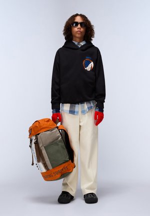 Zwarte hoodie met logo, gelaagd over een blauwe geruite shirt. Crème kleurige broek en rode handschoenen. Multi-kleurige rugzak met oranje accenten aan de zijkant vastgehouden.