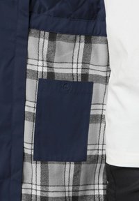 Marineblaue gesteppte Stoff mit einem grauen Karolining und einer dunkelblauen aufgesetzten Tasche; strukturiertes und geschichtetes Design mit kontrastierenden Mustern.