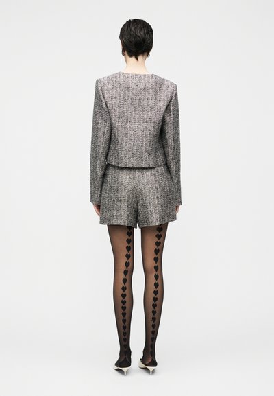 Hallitsetud hallist tweedist bleiser ja lühikeste pükste komplekt, mida täiendab mustade südamemustritega sukkpüksid ja teravate kontsadega kingad, vaadatuna tagantpoolt.