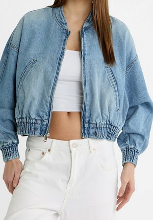 Vrouw draagt een lichtblauw cropped spijkerjasje over een witte cropped top en witte broek met hoge taille, staand tegen een effen achtergrond.