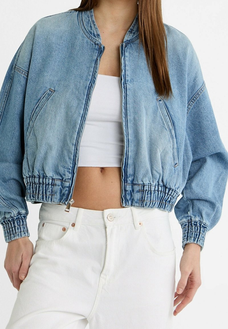 Femme portant une veste courte en denim bleu clair sur un crop top blanc et un pantalon blanc taille haute, debout devant un fond uni.