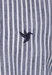 Tejido a rayas en navy y blanco con un acabado texturizado, que presenta un pequeño colibrí navy bordado en el lado izquierdo.