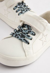 Balti ādas apavi ar velcro siksniņu, kas aprīkoti ar ziliem leopardu raksta šņorēm un perforētu zvaigznīšu apdari sānos.