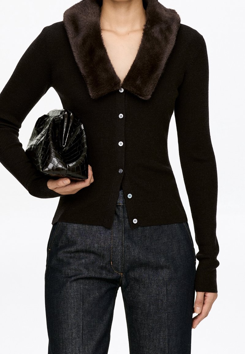 Femme portant un cardigan noir ajusté avec un col en fourrure marron, jean foncé, tenant une pochette noire texturée sous le bras gauche.