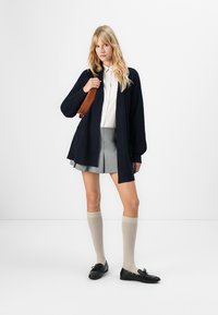 Jeune femme portant une chemise blanche, un cardigan bleu marine, une jupe grise plissée, des chaussettes beige montantes, des mocassins noirs et portant un sac marron à l'épaule.