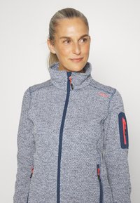 Fleecejack in een blauw-wit visgraatpatroon, met een hoge kraag, marineblauwe accenten en ritszakken met rode hengsels.