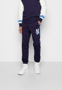 Námořnické tepláky s bílými a modrými akcenty, s vyšitým písmenem "N" a bočními pruhy, vyrobené z měkké, texturované látky.