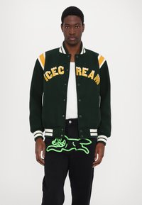 Green varsity jacket with orange and white stripes and "ICE CREAM" in yellow letters. Black pants featuring a neon green animal graphic.