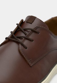 Chaussure à lacets en cuir marron avec finition lisse, coutures visibles et semelle de couleur claire sur fond blanc.