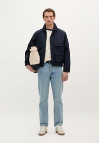 Mørkeblå jakke med to brystlommer, båret over en cremefarvet skjorte. Lys blå jeans og hvide sneakers. Beige taske med en mørkeblå bund.