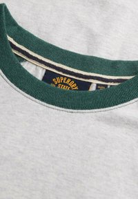 Tæt på en lysegrå sweatshirt-udskæring med en mørkegrøn ribkant og et synligt Superdry-mærke indeni.