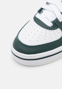 Gros plan sur un bout de basket blanche avec une garniture en cuir vert foncé, des lacets blancs et de petits trous de ventilation sur un fond clair.