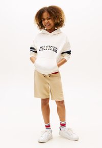Felpa bianca con strisce blu navy e rosse, con logo "HILFIGER". Pantaloni corti beige, sneakers bianche e calze a strisce rosse, bianche e blu.