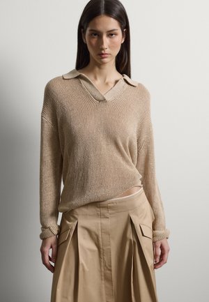 Donna che indossa un maglione beige a maniche lunghe con colletto e una gonna plissettata color tan a vita alta con tasche, in piedi contro un muro semplice.