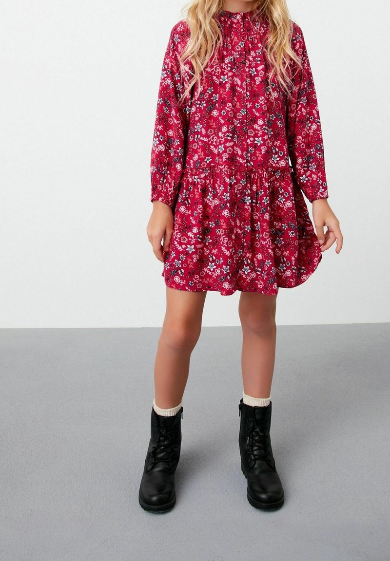 Jeune fille aux longs cheveux blonds portant une robe rouge à fleurs à manches longues et des bottines noires à lacets avec des chaussettes blanches.