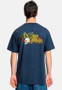 Navy blue cotton t-shirt featuring a large graphic on the back with a skull, roses, and the text “DC Shoe Company” in yellow.