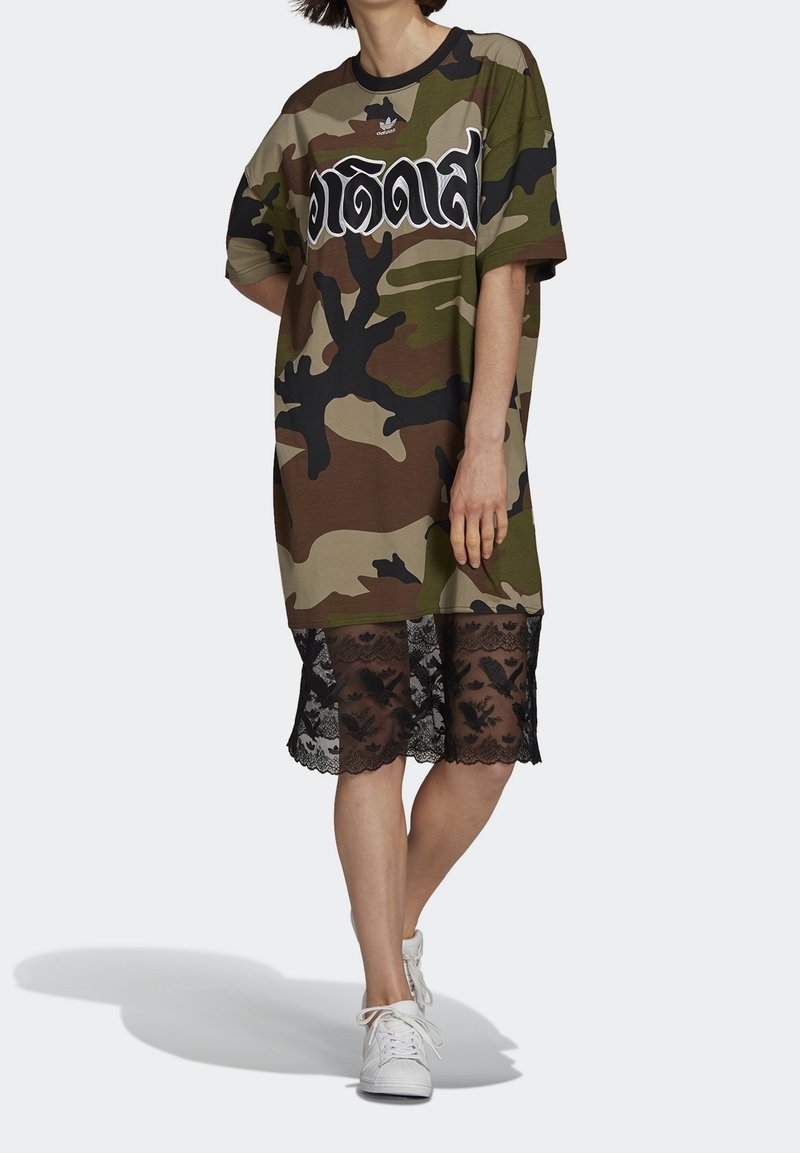 Camo oversized T-shirt in green, brown, and black with white text; layered with black lace trim at the bottom; paired with white sneakers.