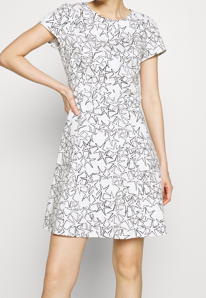 Femme portant une robe blanche à manches courtes avec un motif d'étoile de mer noire, debout avec une main sur la hanche devant un fond blanc.