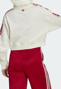 Krátká krémová mikina s kapucí a červeným logem na zádech, kombinovaná s červenými sportovními kalhotami s třemi bílými pruhy po stranách.