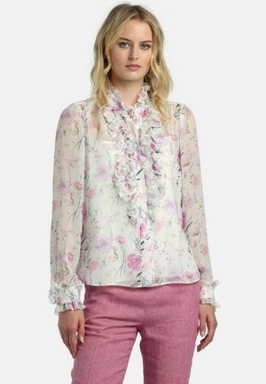 Femeie purtând o bluză albă transparentă cu imprimeu floral roz și volane pe partea din față și la manșete, asortată cu pantaloni roz cu talie înaltă.