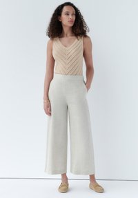 Top tank beige lavorato a maglia con un design a zigzag, abbinato a pantaloni in lino beige chiaro a gamba larga e scarpe slip-on color cammello. Linee semplici e pulite.