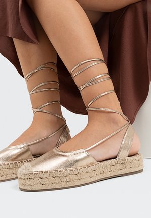 Espadrilles métalliques dorées avec une plateforme en corde de jute, dotées d'une pointe arrondie et de brides croisées à la cheville pour un maintien sécurisé.