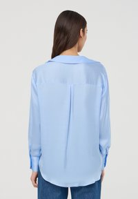 Blusa azzurro chiaro con una texture liscia e setosa, maniche lunghe e colletto pieghettato. Presenta una piega sul retro e un orlo leggermente curvo.