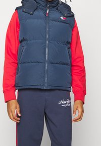 Granatowa pikowana kamizelka z zamkiem z przodu i odpinanym kapturem, noszona na czerwonej koszulce z długim rękawem, w połączeniu z granatowymi dresami z logo.