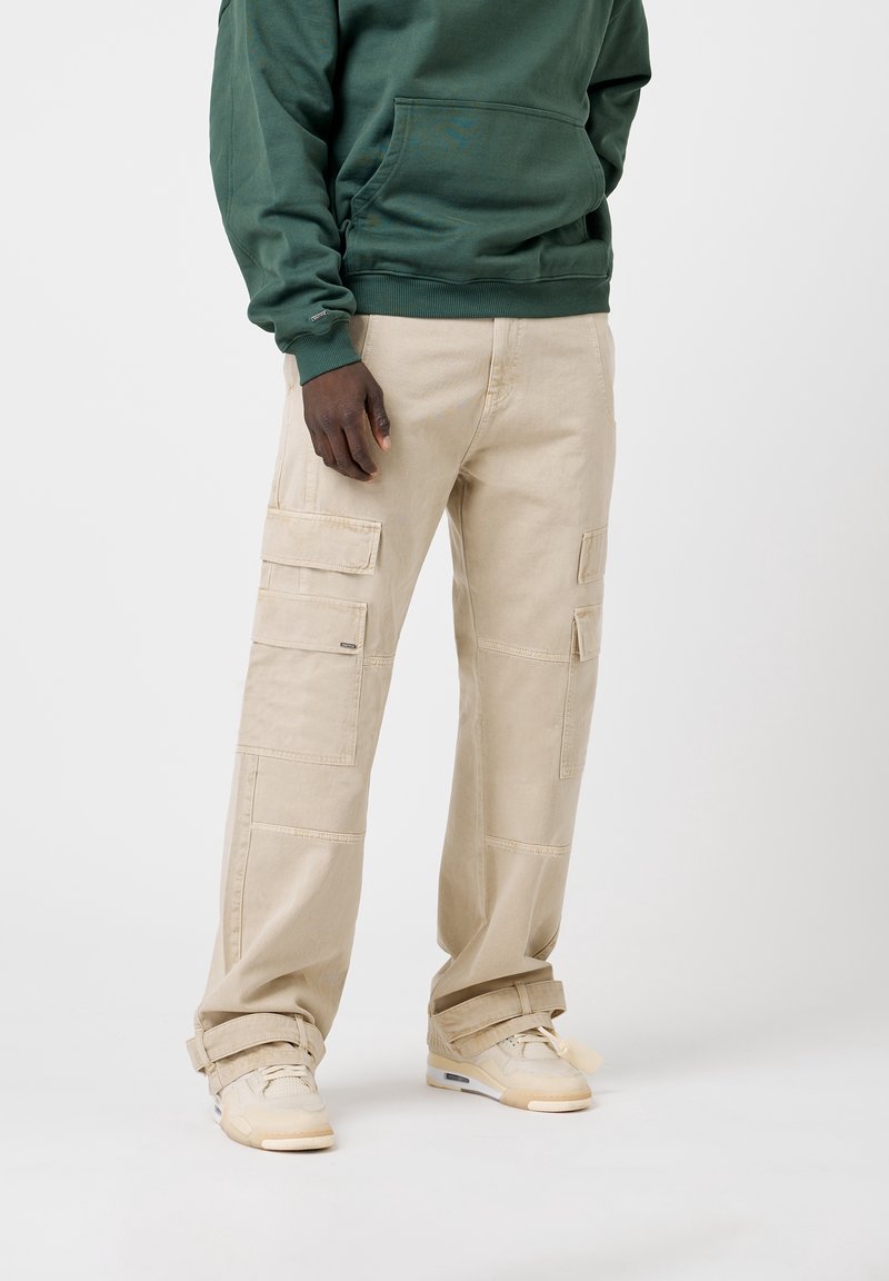 Pantalon cargo beige en coton avec multiples poches, coupe droite et coupe décontractée, assorti à des baskets de couleur claire.