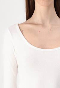 Balta ilga rankovė su apvaliu kaklu, turinti išraizgytą tekstūrą ir prigludusį dizainą. Mažas pusmėnulio tatuiruotė matoma šalia raktikaulio.
