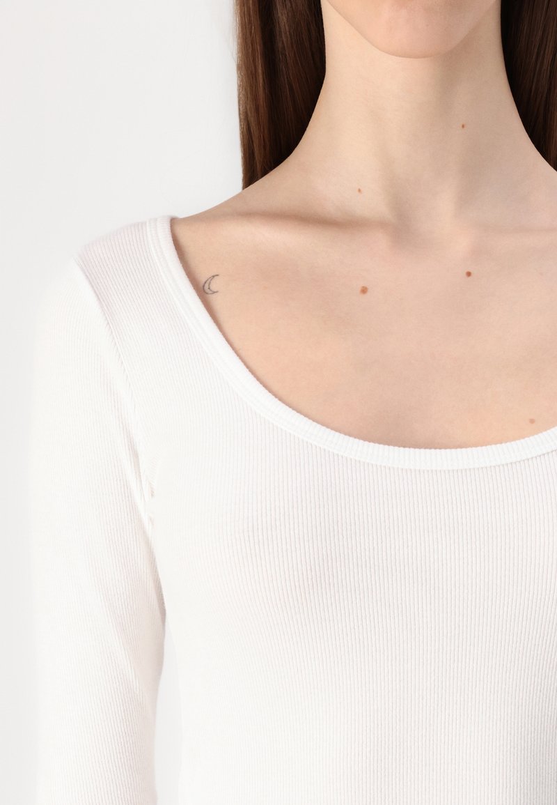 Weißes Langarmoberteil mit Rundhalsausschnitt, ausgestattet mit einem gerippten Design und figurbetonter Passform. Kleines Halbmond-Tattoo in der Nähe des Schlüsselbeins sichtbar.