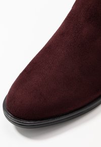 Bottine en daim bordeaux à texture lisse, bout arrondi et semelle en caoutchouc noire. Le design est épuré et minimaliste.