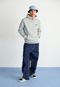 Felpa grigia con cappuccio, pantaloni di denim scuri e oversize, cappello a secchiello blu, sneaker nere e gialle. Sfondo semplice, pavimento blu chiaro.
