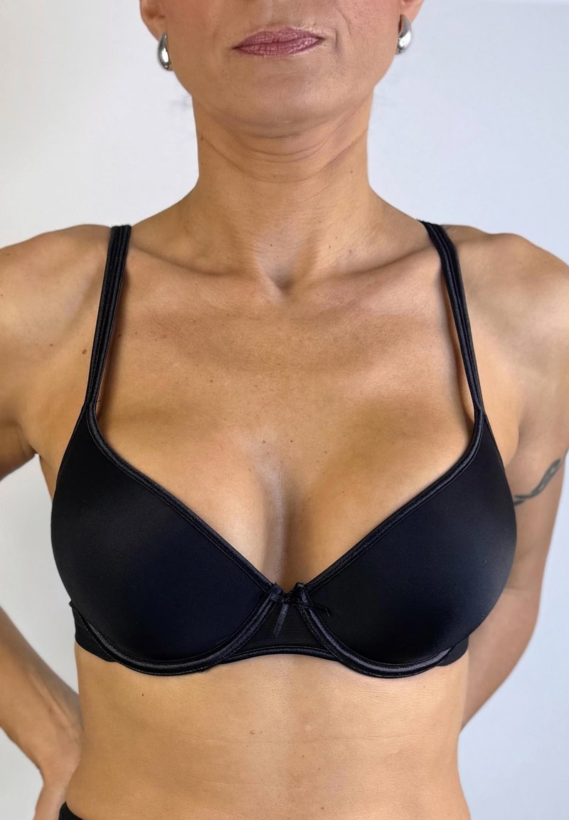 Sujetador negro acolchado con tirantes finos ajustables, con un escote en forma de V y un pequeño lazo decorativo en el centro. Textura de tela suave.