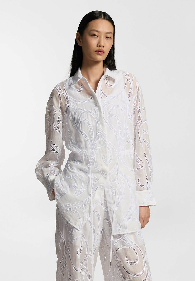 Femme aux longs cheveux noirs portant un chemisier blanc transparent et un pantalon assorti avec des motifs brodés en spirale, posant les mains dans les poches.