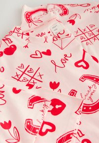 Lyserødt stof med røde trykte mønstre, herunder hjerter, blomster og fraser som "love" og "lucky you." Rund halsudskæring, blød tekstur.