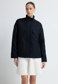 Granatowa teksturowana kurtka z wysokim kołnierzem, dwoma przednimi kieszeniami i szerokimi rękawami, w zestawie z białą wełnianą spódnicą.