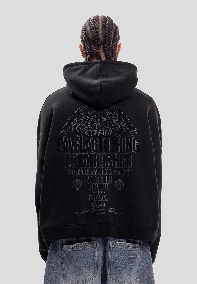 Personne aux cheveux tressés porte un sweat à capuche noir avec un texte en gras "FAVELACLOTHING ESTABLISHED" et un jean bleu délavé avec de grandes poches à l'arrière.