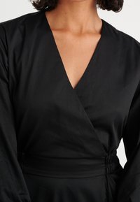 Schwarzes Wickelkleid mit tiefem V-Ausschnitt, elastischem Bund und langen Ärmeln. Der Stoff wirkt glatt mit matter Oberfläche.