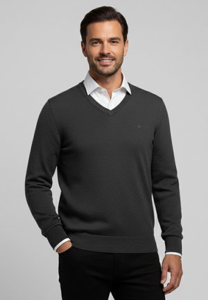 Hombre sonriente, vistiendo un suéter de cuello en V gris oscuro sobre una camisa blanca con cuello, con una mano en el bolsillo, contra un fondo gris liso.