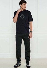 T-shirt nera in cotone con maniche corte e una grafica a forma di diamante sul petto, abbinata a pantaloni scuri e sneakers nere con dettagli bianchi.