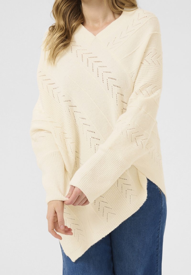 Krämfärgad stickad poncho med v-ringning, som har ett zigzag-mönster och ribbade detaljer. Designen har en asymmetrisk fåll och en lös passform.