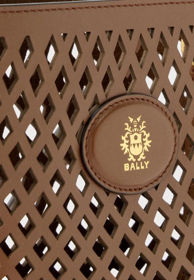 Κοντινό πλάνο καφέ δέρματος με τρυπήματα σε σχήμα ρόμβου και κυκλικό επίθεμα με ανάγλυφο το λογότυπο της Bally σε χρυσό.