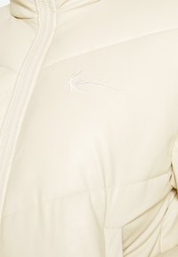 Cream quilted jacket with zip closure and high collar. Features subtle logo embroidery and smooth, shiny synthetic material.