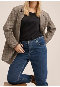 Blazer à carreaux marron et noir porté par-dessus un haut noir, associé à un jean bleu foncé. Vue en gros plan mettant en valeur les motifs du tissu et la coupe.