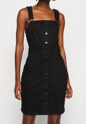Sort denim overallskjole med skulderstropper, firkantet udskæring, knaplukning foran og to store lommer. Glat tekstur og skræddersyet pasform.