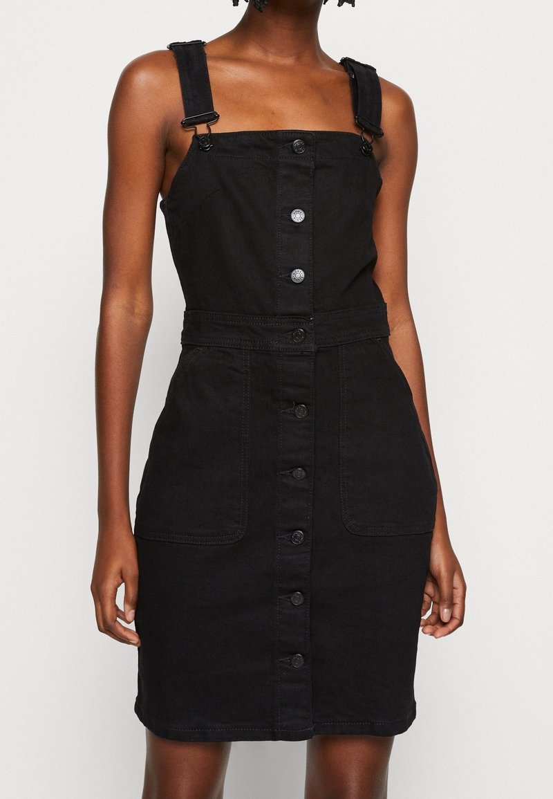 Robe salopette en denim noir avec bretelles, encolure carrée, fermeture à boutons sur le devant et deux grandes poches. Texture lisse et coupe ajustée.