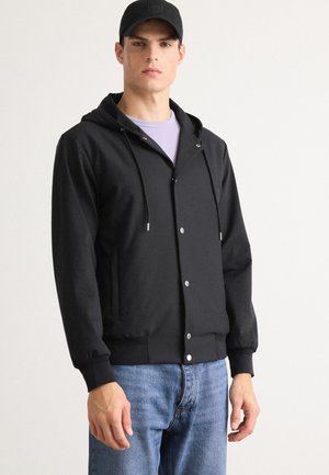 Un jeune homme portant une veste à capuche noire à boutons-pression, une chemise violet clair en dessous, une casquette noire et un jean bleu, se tenant contre un fond blanc.